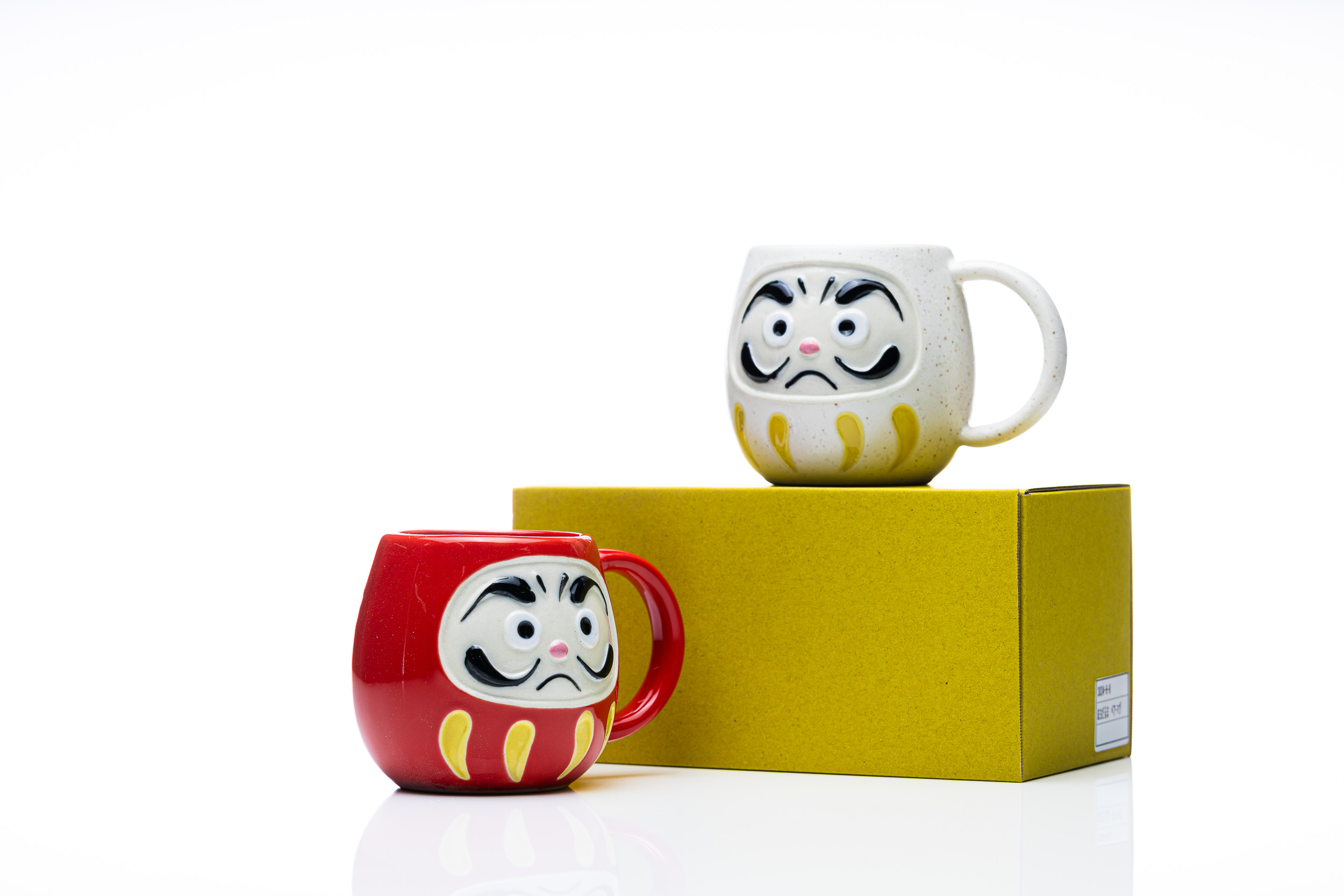 Two daruma mugs with a yellow product box on a white studio background